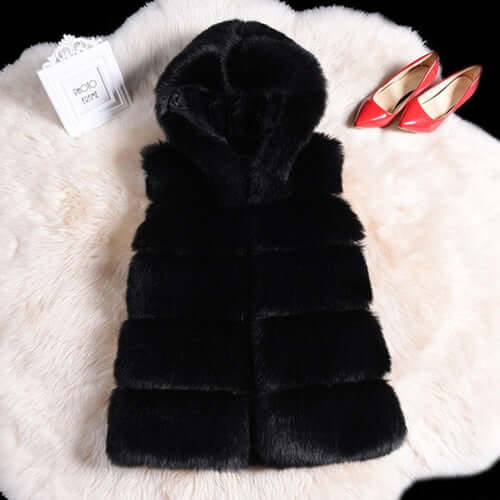 Black sleeveless faux fur coat with hood on a fluffy backdrop, paired with red high heels and a decorative box.