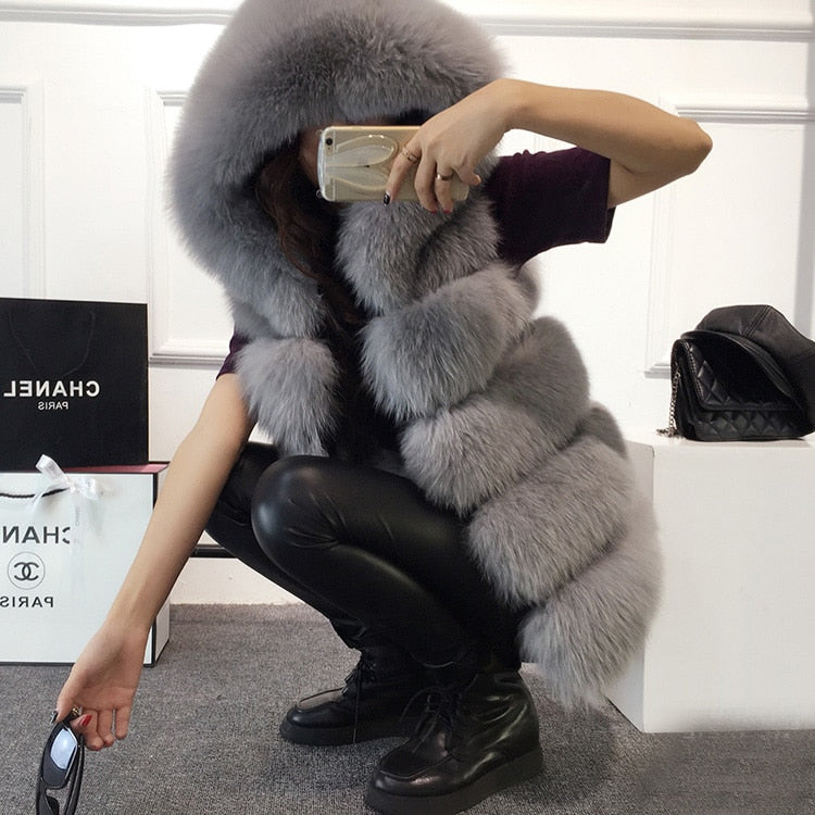 Woman in stylish 2022 winter sleeveless artificial fox fur coat gilet and leather pants, covering face with phone, sitting indoors.