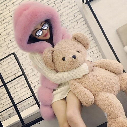 Woman wearing a pink hooded faux fur coat hugging a large teddy bear in a cozy indoor setting.