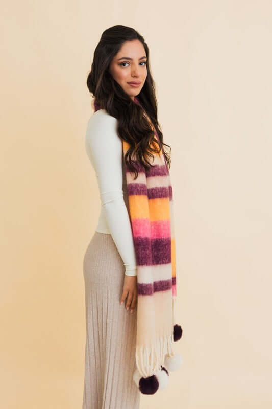 Woman wearing a contrast color striped chunky pom tassel scarf against a light background, showcasing bold patterns and playful design.