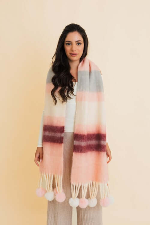 Woman wearing a chunky pom tassel scarf with contrast color stripes in shades of pink, gray, and burgundy against a light background.