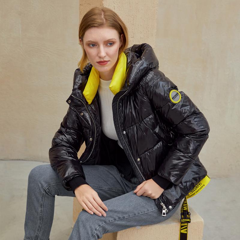 Woman wearing short black winter coat parka with hood, paired with gray jeans and a bright yellow scarf, sitting casually.