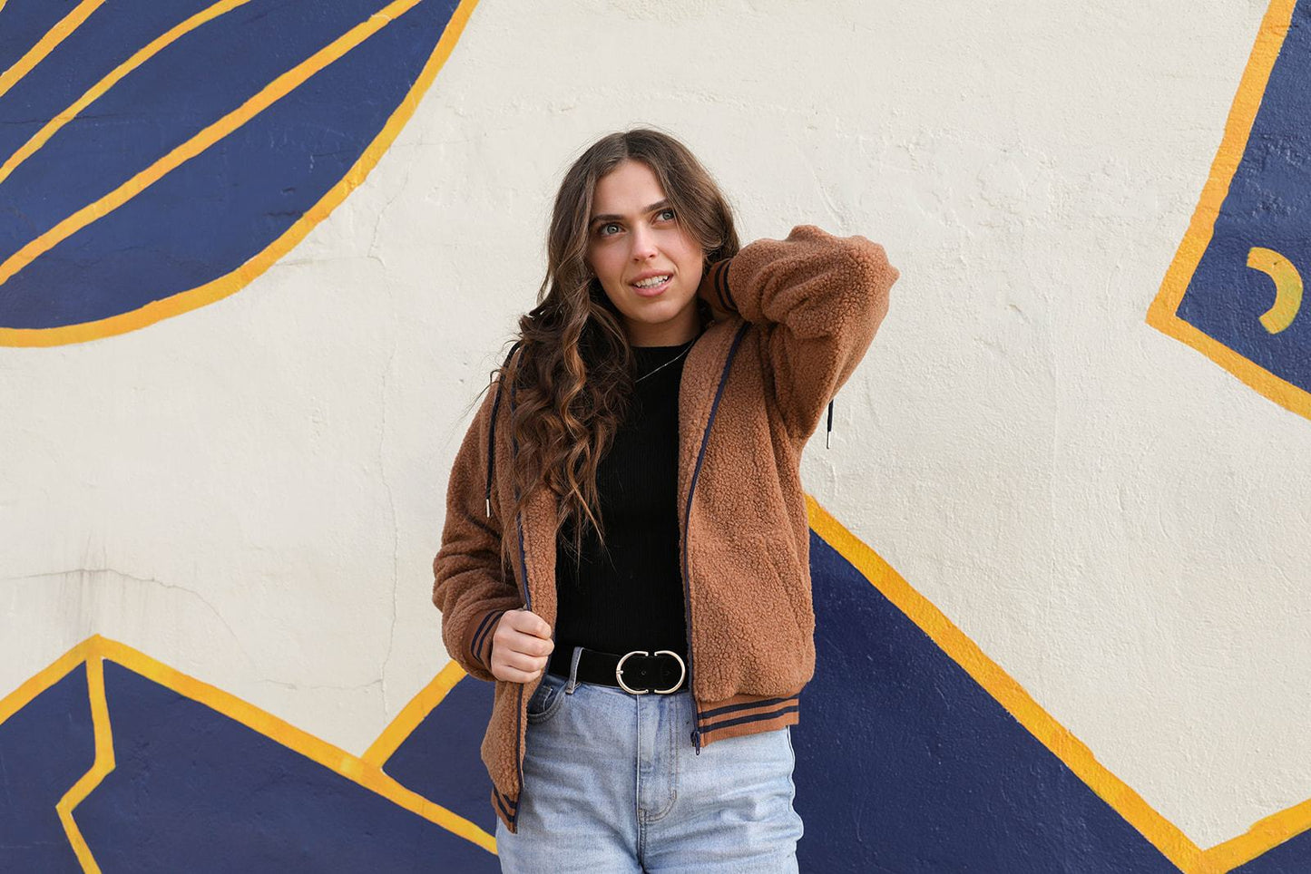 Woman in brown DT Bode Sherpa winter jacket and jeans standing against a patterned wall, exuding a cozy and fashionable vibe.