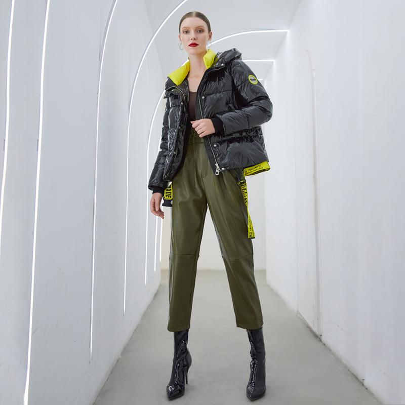 Woman in a modern hallway wearing a short shiny winter coat with hood and olive green pants, styled for a contemporary look.