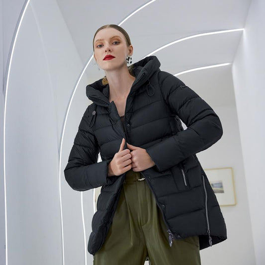 Woman modeling a black hooded winter coat with full sleeves in a modern white curved interior setting.