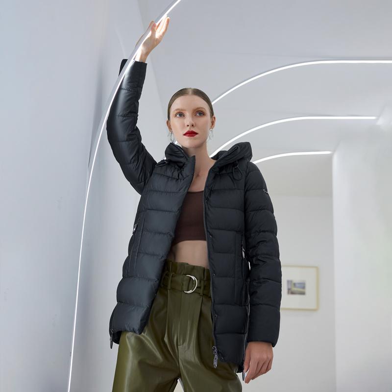 Woman wearing a black hooded winter coat and green trousers, posing in a modern room with curved light panels