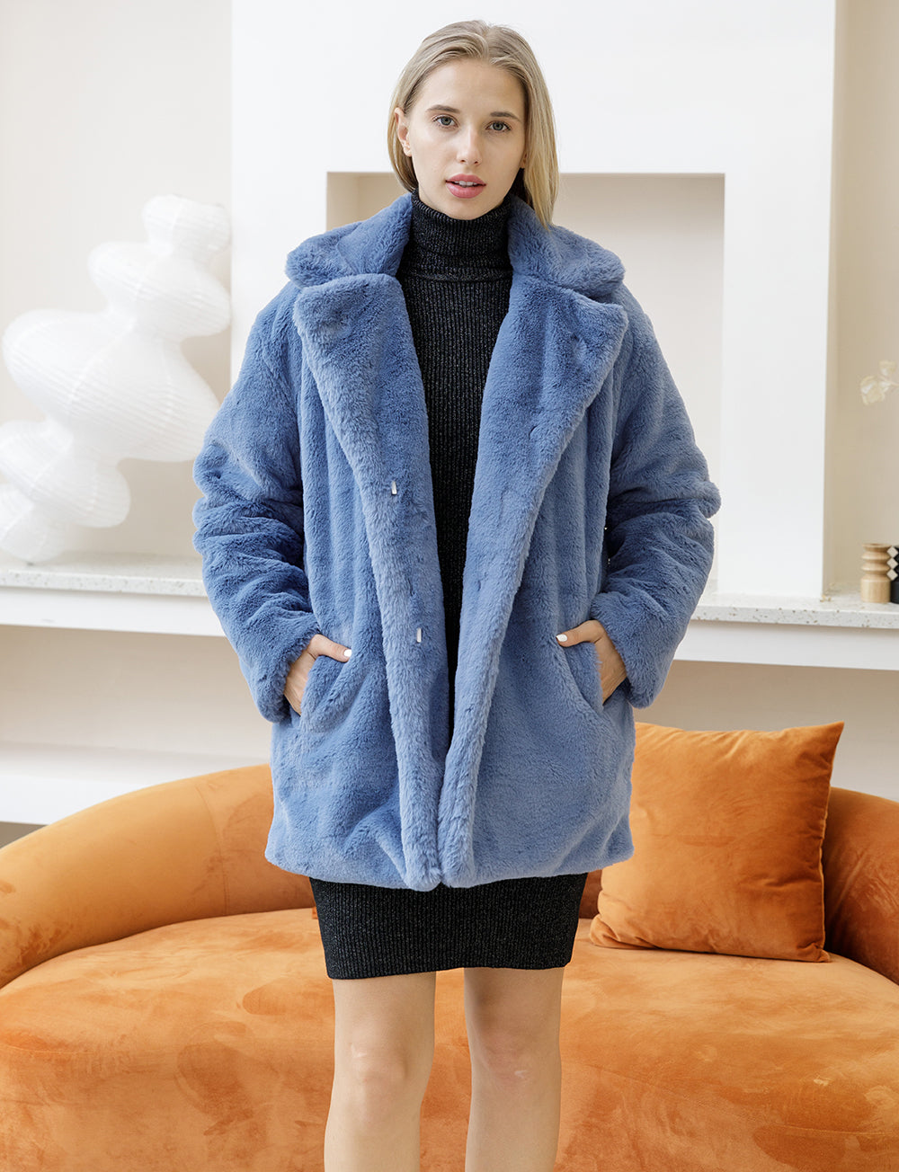 Woman modeling 2022 Autumn Winter faux fur coat in blue, featuring plush texture and elegant turn-down collar, indoors near orange sofa.