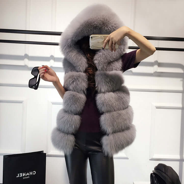 Stylish woman wearing a sleeveless faux fur hooded gilet in gray, holding sunglasses, showcasing winter outerwear fashion.