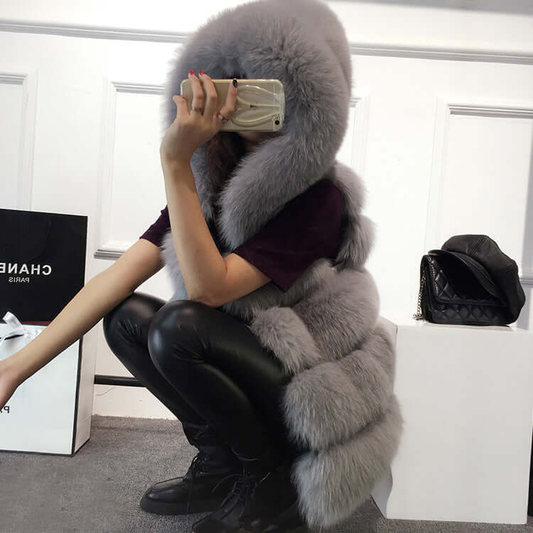 Woman wearing a gray sleeveless faux fur coat with a hood, posing indoors with stylish accessories.