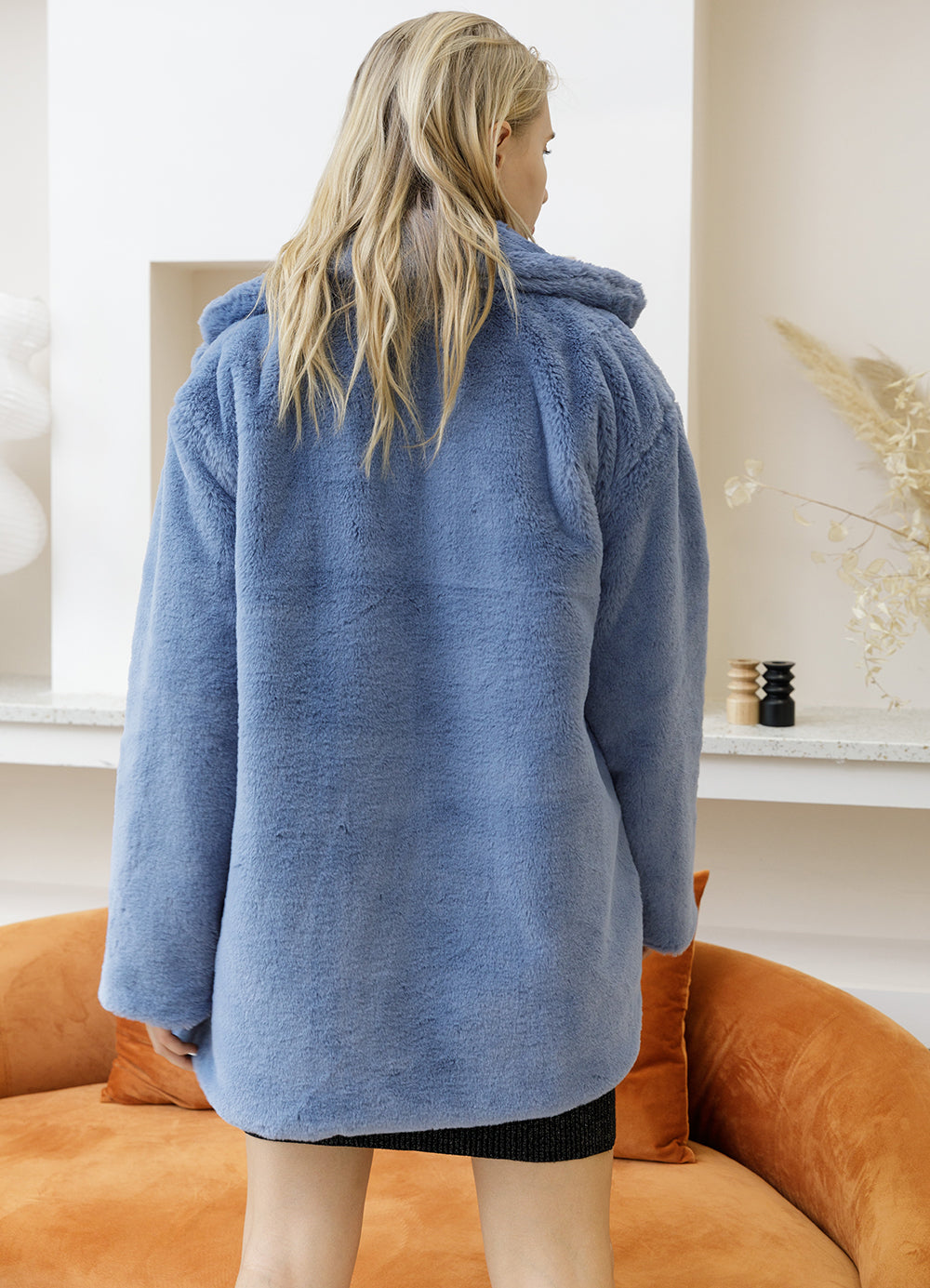 Woman wearing a 2022 blue faux fur coat with turn-down collar, viewed from behind in a modern living room setting.