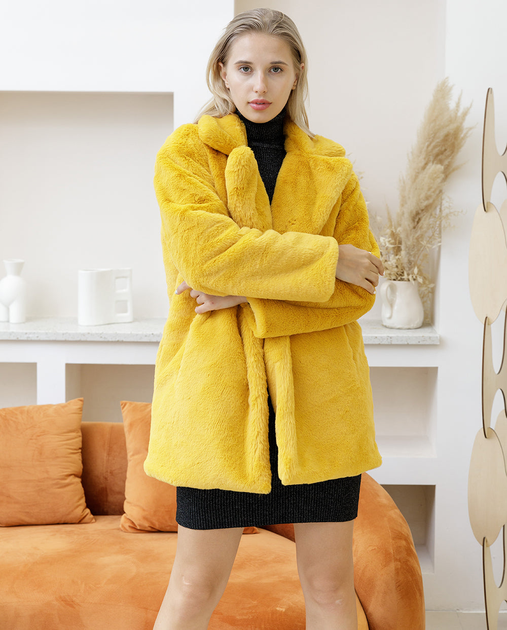 Woman wearing a yellow faux fur coat from the 2022 Autumn Winter collection in a stylish interior setup with orange sofa and decorative items.