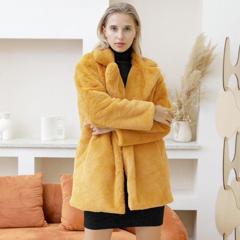 Woman wearing a 2022 Autumn Winter faux fur coat in elegant yellow, standing in a stylish living room.