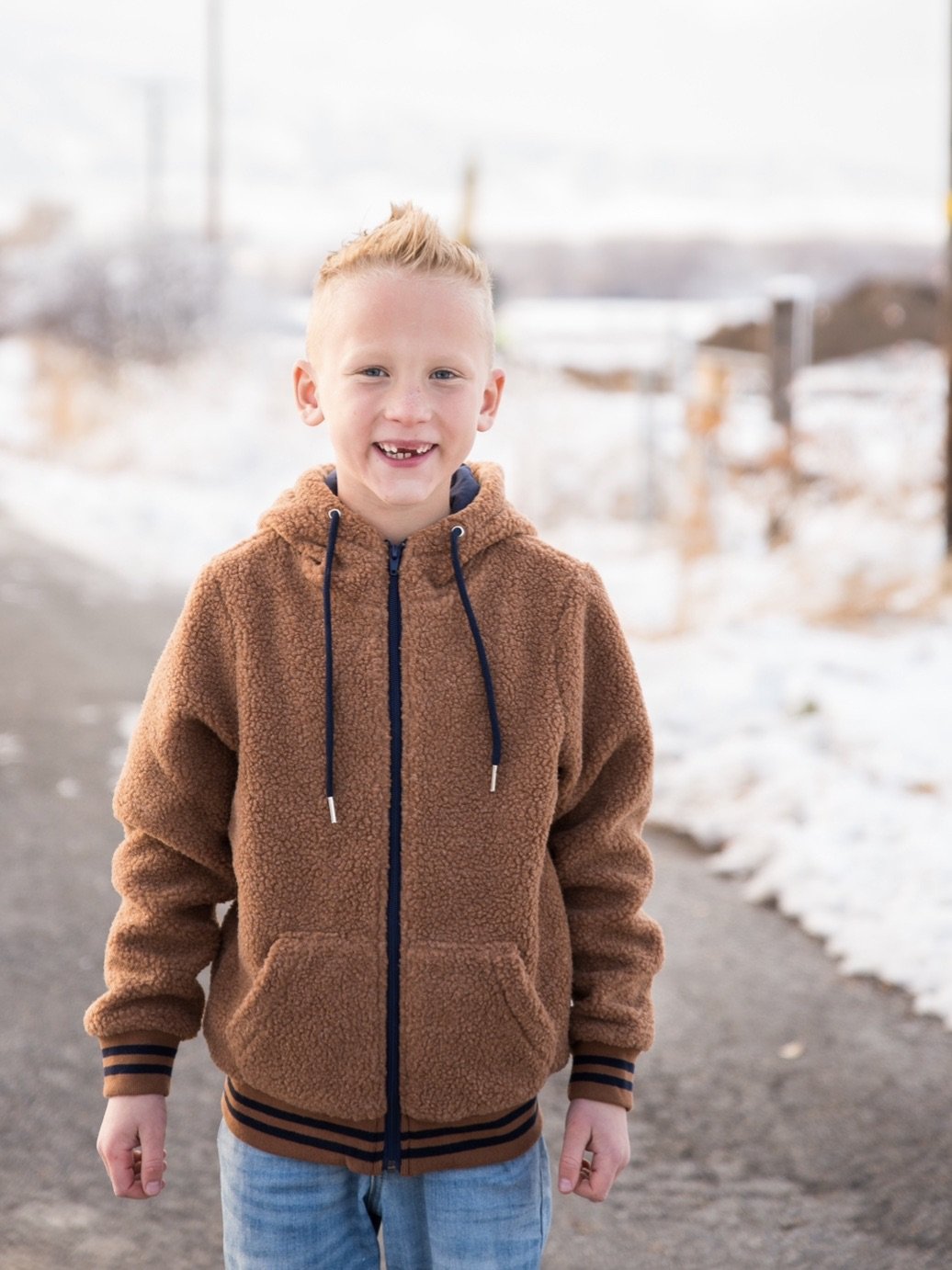 Young boy smiling in a brown sherpa winter jacket with blue accents, outdoors on a snowy path.