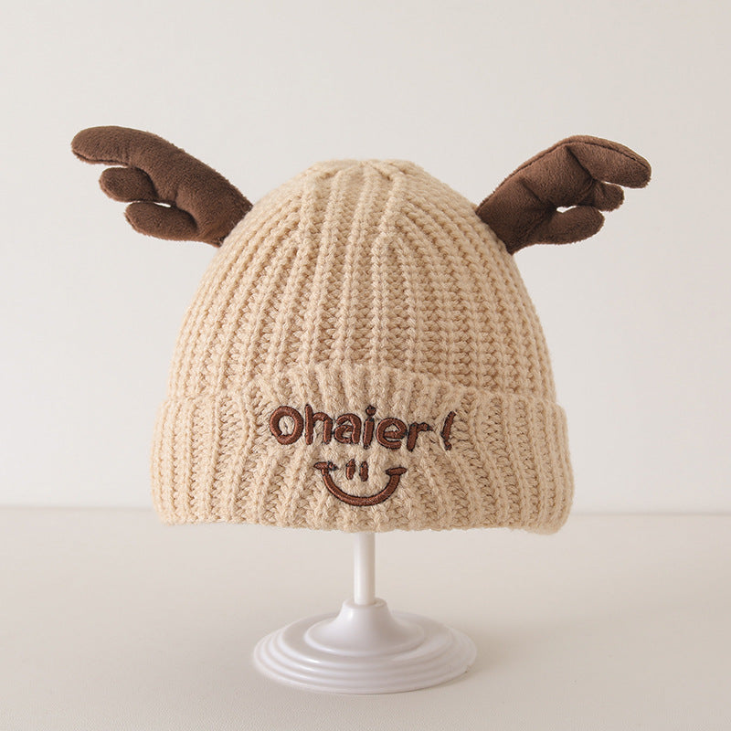 Cute beige baby knit hat with embroidered smile and brown deer antlers displayed on a white stand.