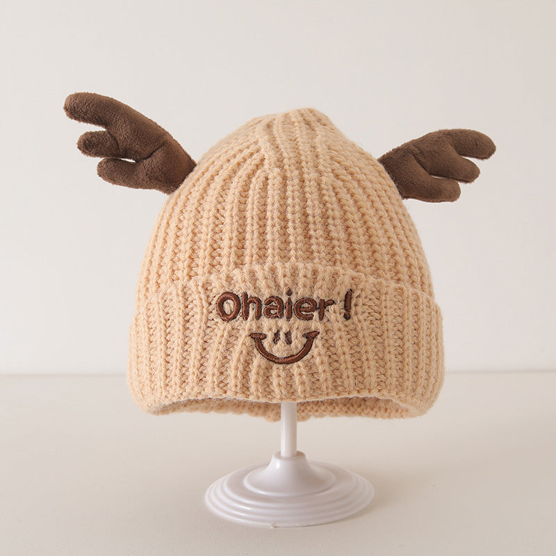 Beige knit baby hat with cute deer antlers and embroidered smiling face on display stand, ideal for winter and autumn