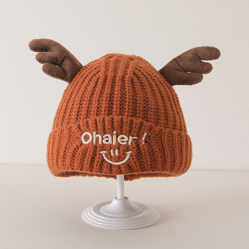 Baby's orange knit hat with cute deer antlers and embroidered smile displayed on a stand, perfect for autumn and winter styles.