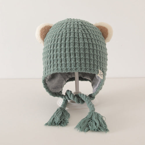 Cute green knitted baby balaclava with bear ears and braided tassels on a display stand, cozy winter and autumn accessory.