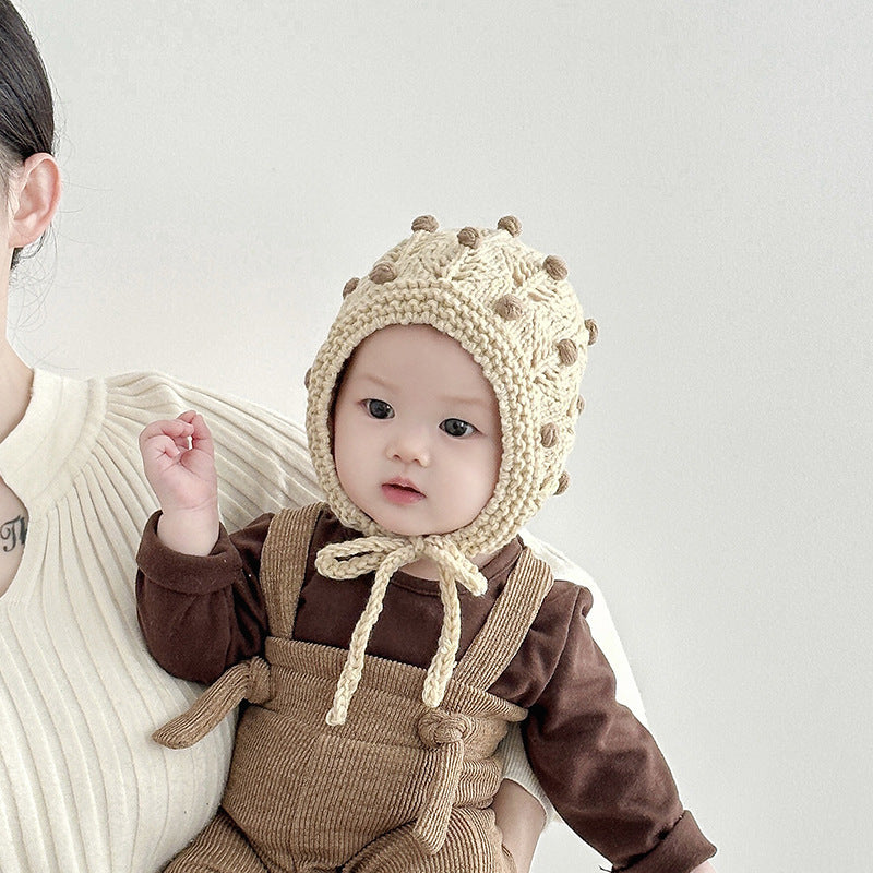 Baby in beige vintage design warm balaclava with dot pattern, wearing a brown outfit, held by parent in cream top.