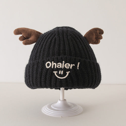 Black knit baby hat with cute deer antlers and embroidered smiley face, displayed on a white stand.