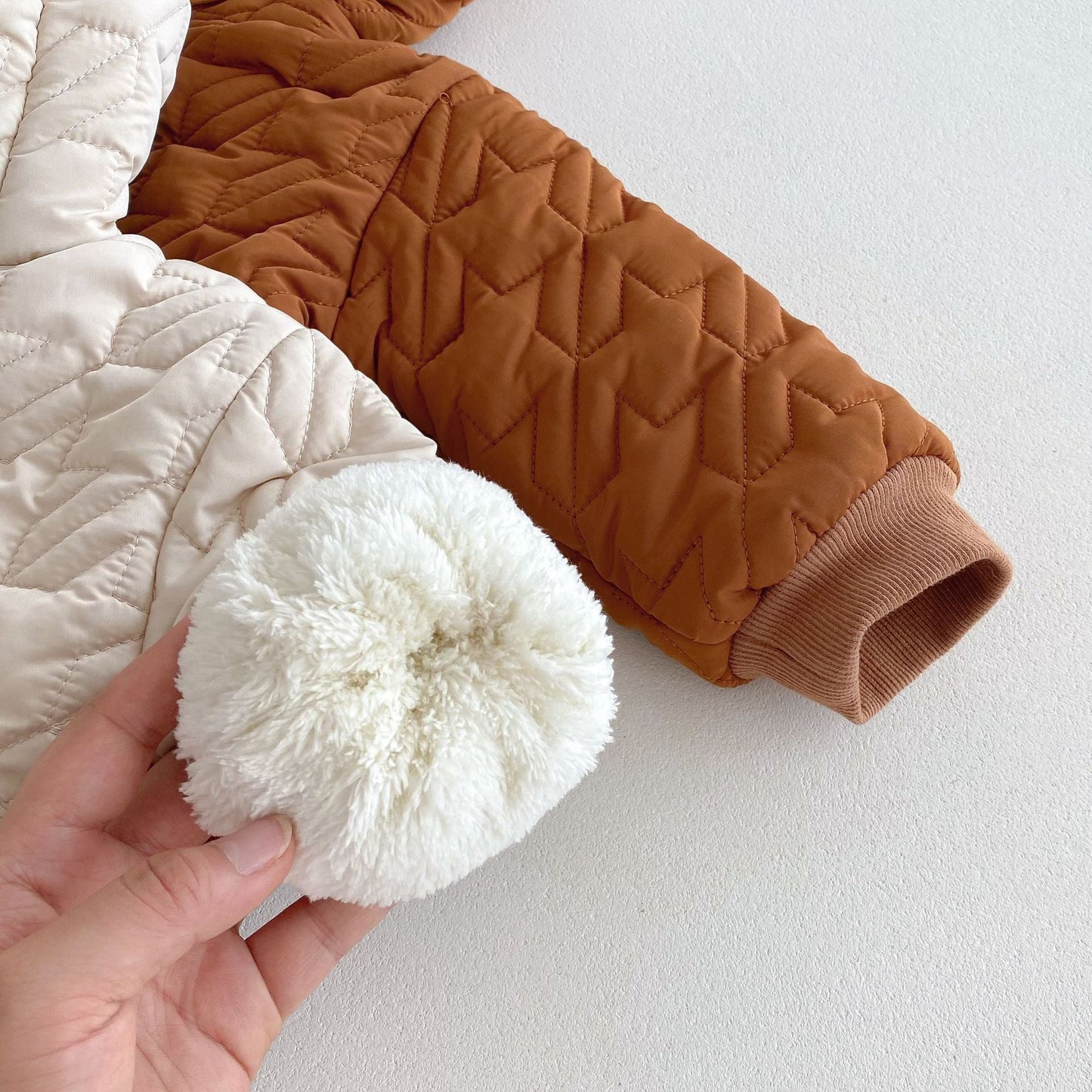 Close-up of a beige and brown quilted infant coat with a fluffy white pom-pom held by a hand