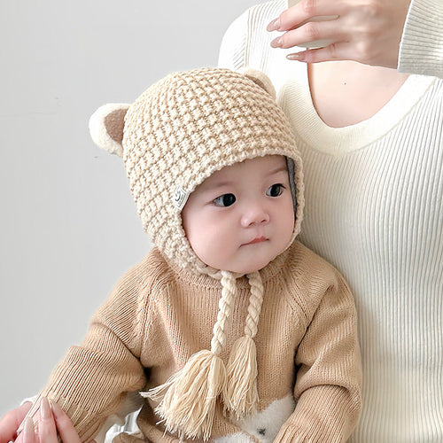 Baby wearing a beige plaid balaclava with cute bear ears, seated next to mother in cream sweater, looking aside.