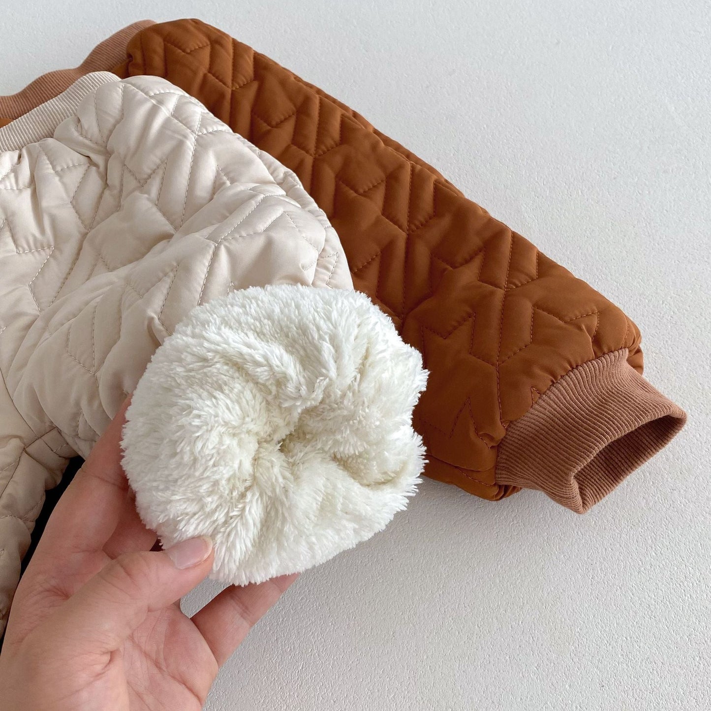 Hand holding a fluffy white cuff of a child's coat next to folded beige and brown quilted pants